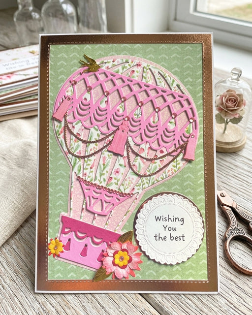 A handmade greeting card featuring a pink and green hot air balloon design with the text 'Wishing You the best' on a scalloped white circle, displayed on a rustic wooden table with a linen cloth and scissors nearby.