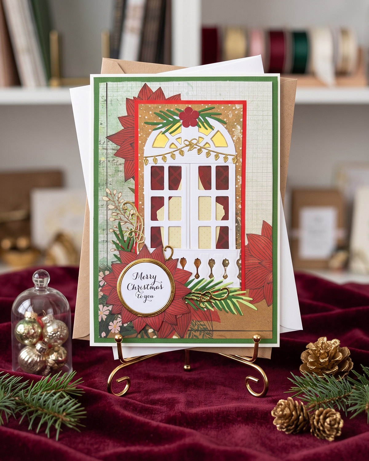 A handmade Christmas greeting card displayed on a small gold stand, surrounded by holiday props including pine branches, gold pinecones, and a glass dome of gold ornaments on a dark red velvet surface. The card features a white window illustration with red poinsettia accents and a circular label reading 'Merry Christmas & you'. Made in NZ by Del Cards NZ
