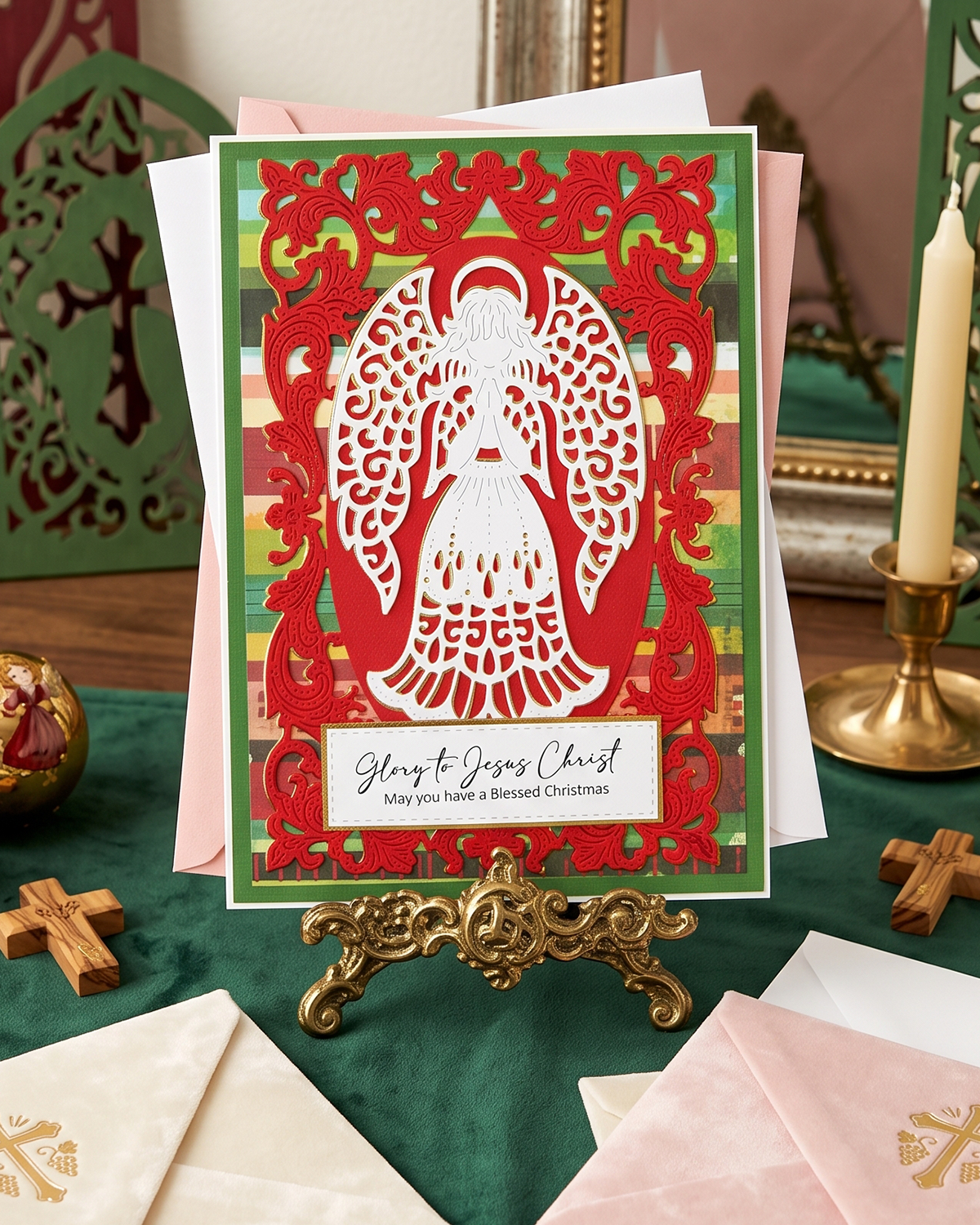 A handmade Christmas greeting card featuring a white angel silhouette cutout over a red ornate border and a green-and-red plaid background, displayed on a gold stand on a dark green table with pink envelopes and candles around it.