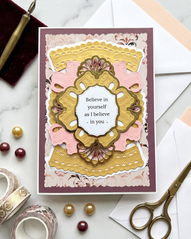 Handmade plum and gold "Believe in yourself" card displayed upright in a styled flat-lay setting with scissors, gold beads and ribbon alongside. Rich plum, blush and antique gold tones make this an elegant handmade card for birthdays, graduations or moments of encouragement, made by Del Cards NZ.