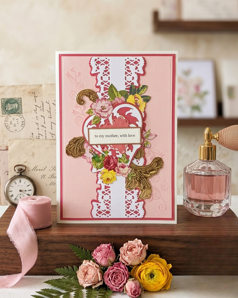 Handmade luxury greeting card with a blush pink background layered on a deep rose cardstock mat. A central white lace-style die-cut column runs vertically, flanked by clusters of hand-applied paper flowers in pink, red and yellow roses with green leaves. A decorative die-cut heart frame sits at the centre, holding a sentiment tag reading "to my mother, with love" in elegant italic script. Gold embossed botanical accents — a leaf spray and a butterfly — add warmth and dime