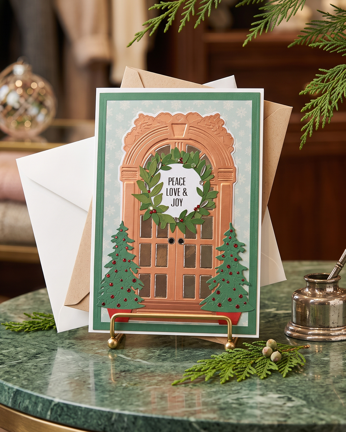 A festive handmade holiday greeting card displayed on a small gold stand on a green marble table, surrounded by pine branches, a white envelope, and a silver inkwell.