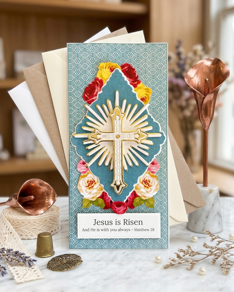 Handmade luxury Easter card displayed upright on a wooden surface beside dried florals and a small decorative object, showing full card design with gold cross, sunburst and rose details, made in NZ by Del Cards NZ