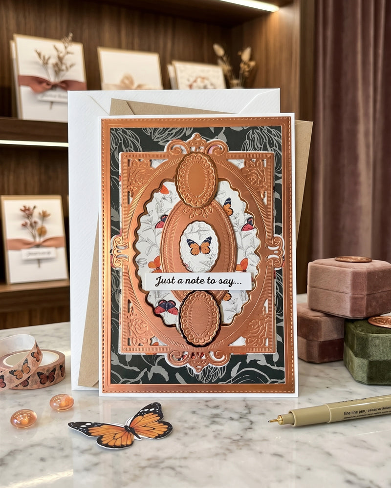 Handmade copper and charcoal "Just a note to say" card displayed upright in a warm styled interior setting with soft props alongside. Rich copper tones against a dark background create a dramatic, elegant handmade greeting card made in New Zealand by Del Cards NZ.