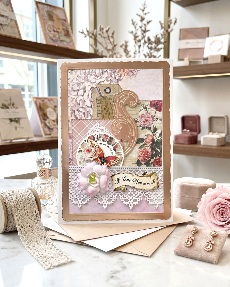 Handmade luxury love card displayed upright on a bright workspace surface beside pink roses and craft materials, showing full card design with gold scroll, lace and vintage floral layers, made by Del Cards NZ