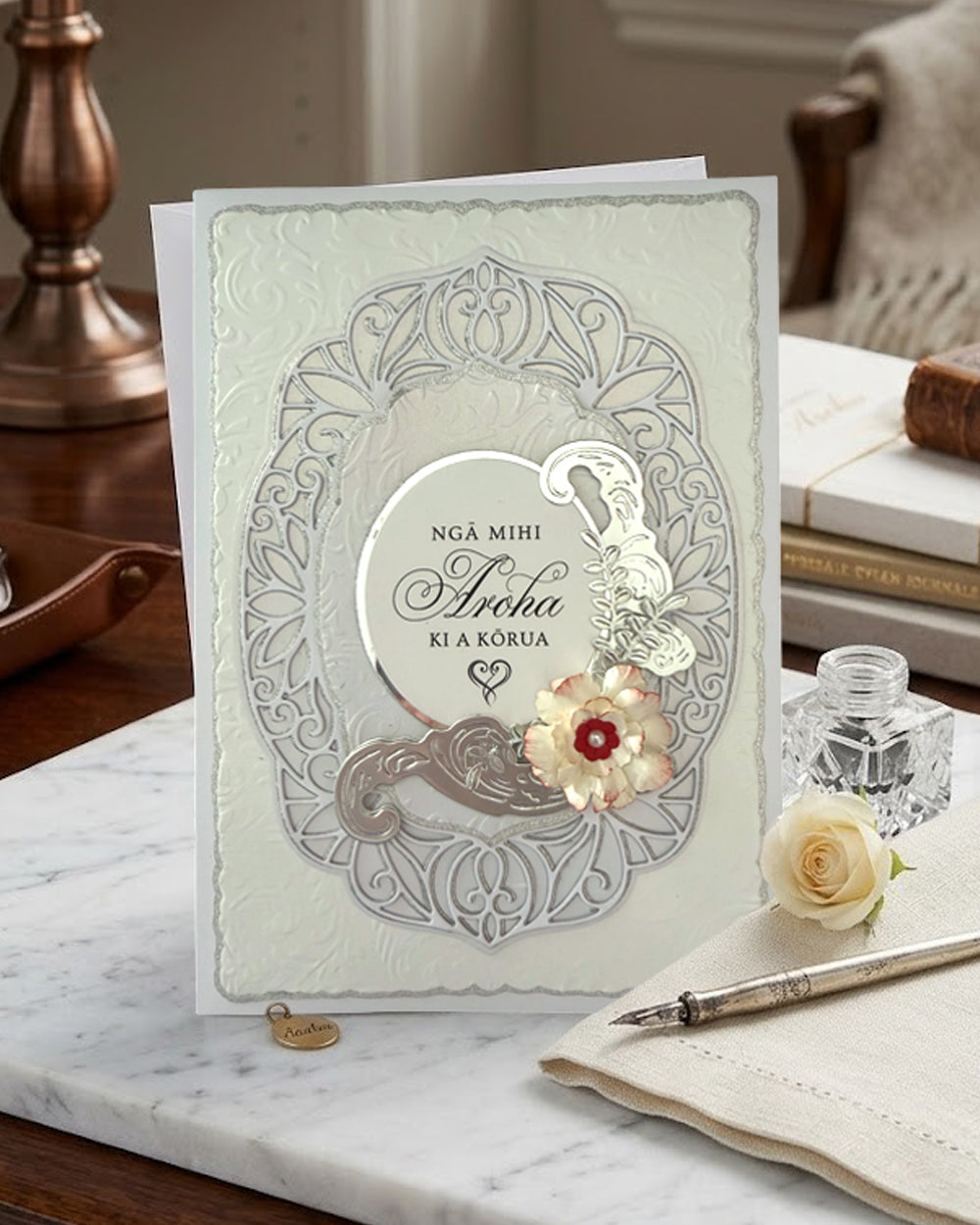 Te Reo Māori Wedding Card displayed upright on a wooden desk beside an open notebook and pen, for sale, made by Del Cards NZ