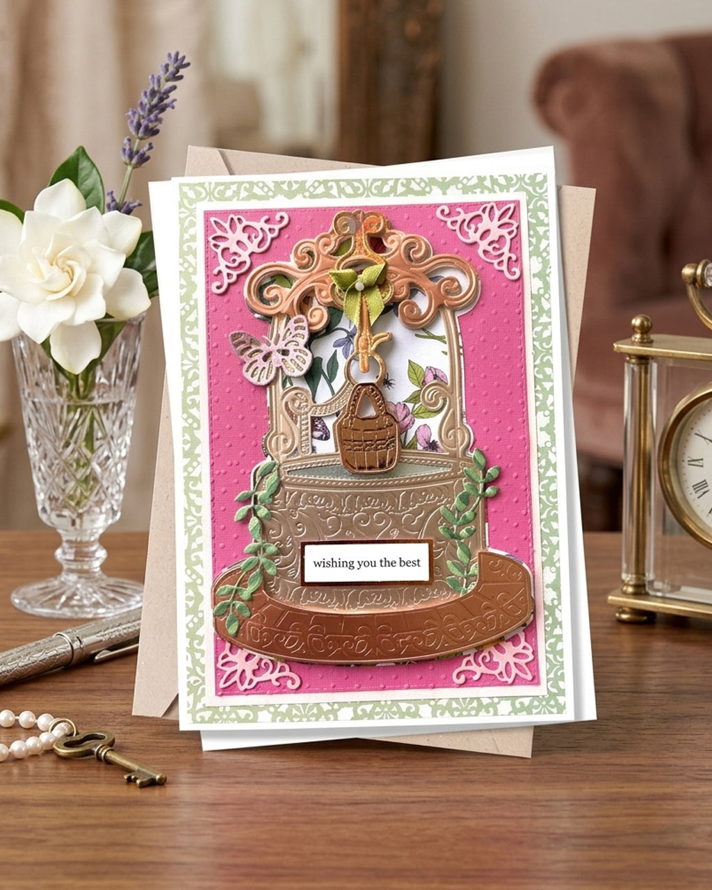 An ornate handmade greeting card displayed on a wooden table, featuring a pink background, a 3D floral arch, a small basket, and a white card that reads 'wishing you the best'. The card is styled with a vase of white flowers, a pen, keys, and a brass clock in the background. Handmade luxury card made by Del Cards in NZ.