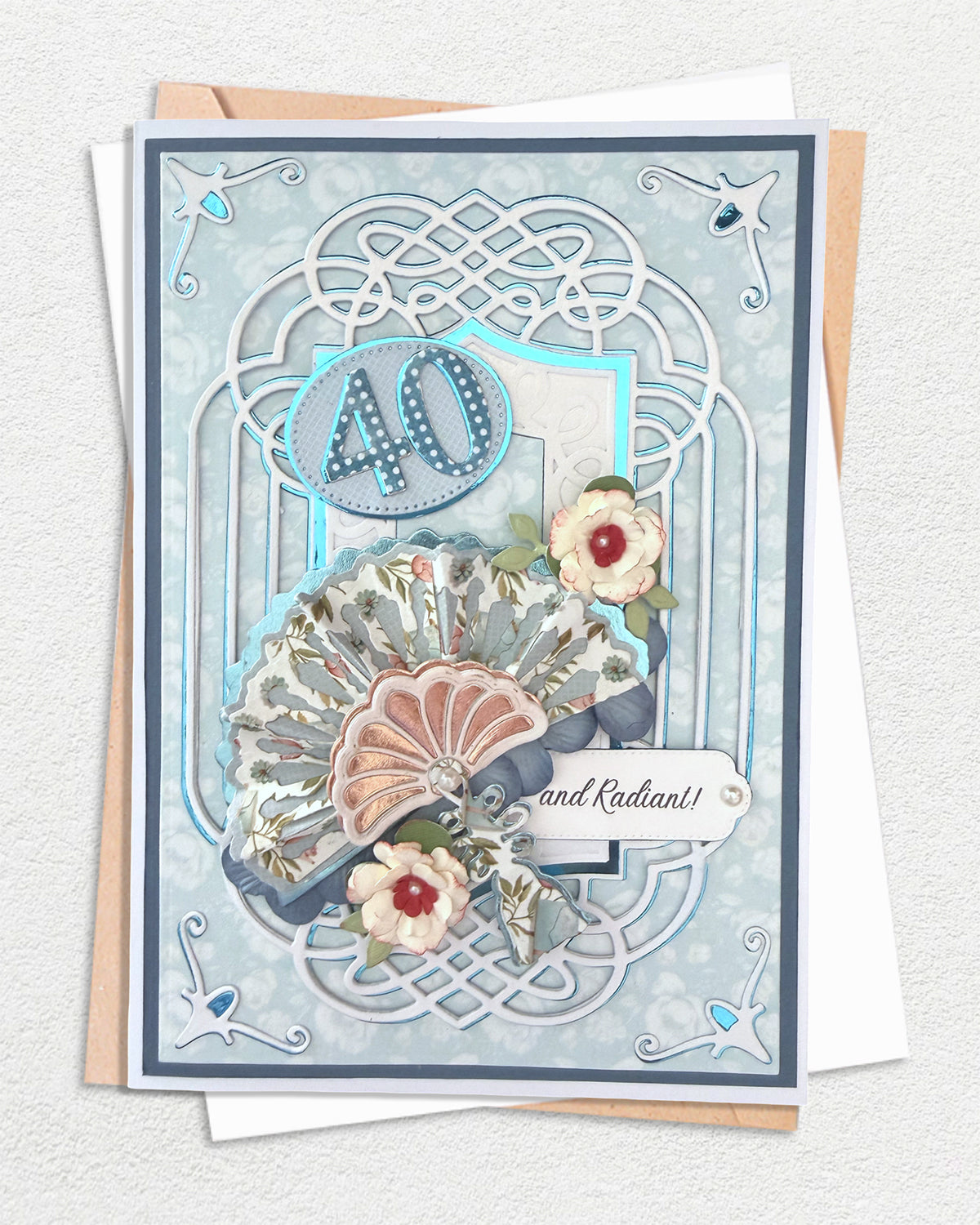 Handmade 40th birthday card with dusty blue Celtic knotwork die-cut frame, silver foil star corner accents, layered paper fans in floral and rose-gold foil, and hand-tied paper roses, with kraft envelope behind.