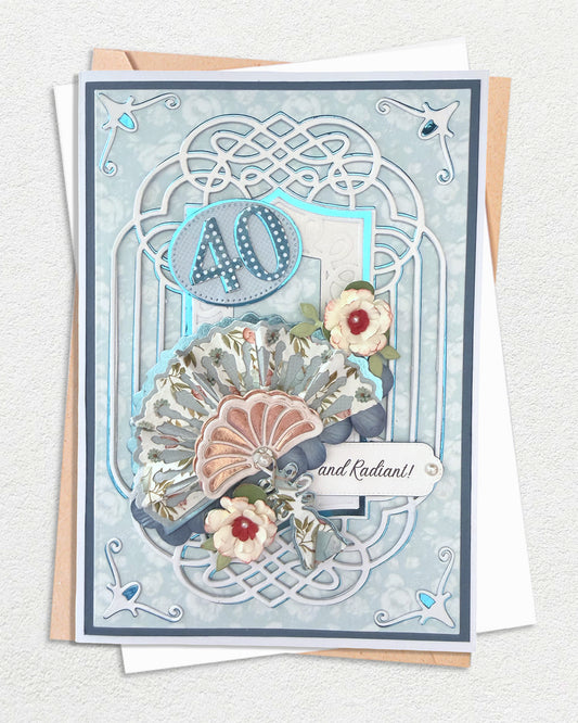 Handmade 40th birthday card with dusty blue Celtic knotwork die-cut frame, silver foil star corner accents, layered paper fans in floral and rose-gold foil, and hand-tied paper roses, with kraft envelope behind.