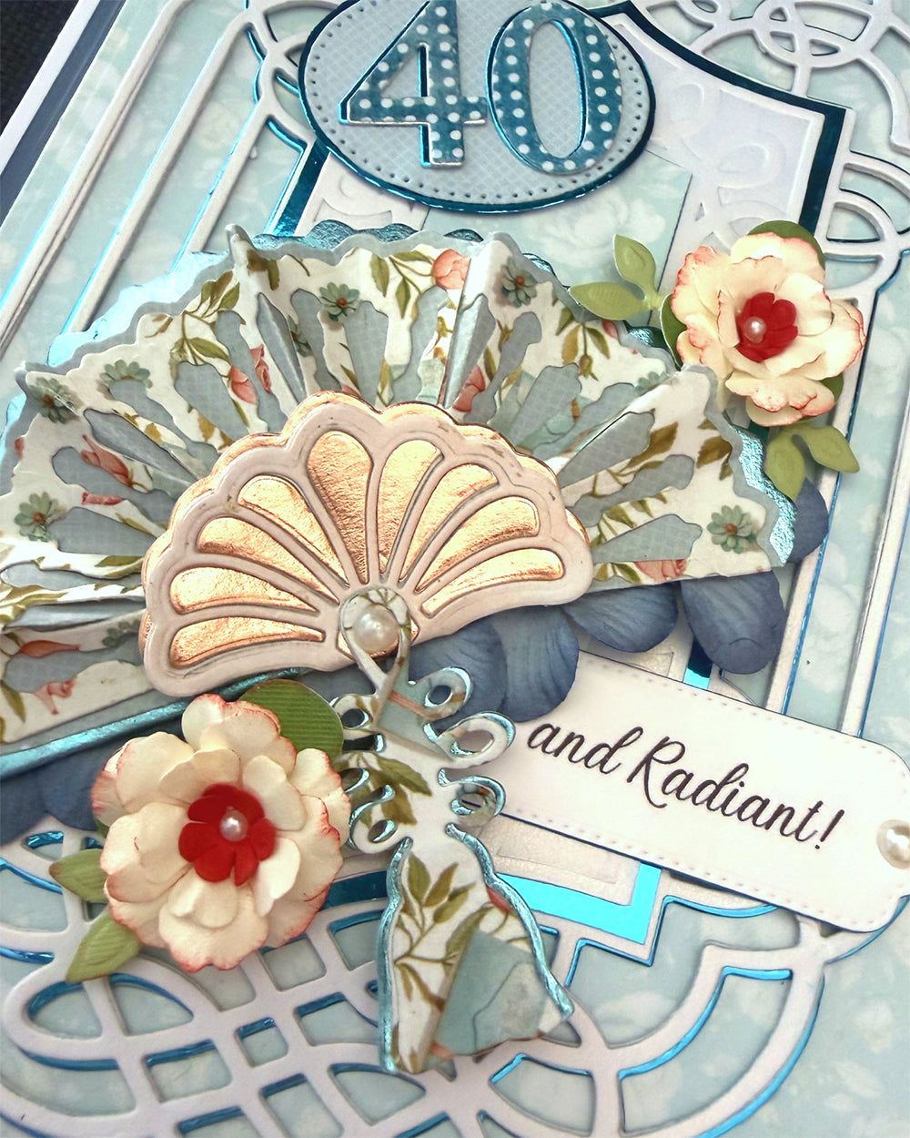 Close-up of "and Radiant!" sentiment tag with pearl accent, beside layered paper fans and hand-tied flowers on a handmade 40th birthday card.