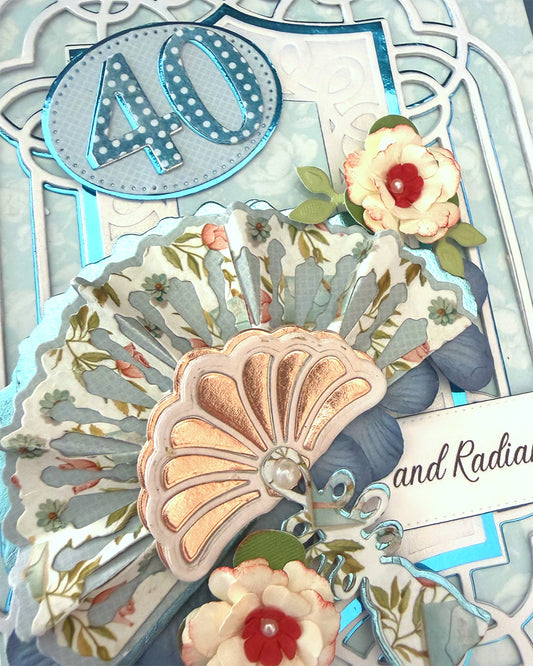 Close-up of hand-folded paper fans in floral patterned paper and rose-gold foil, layered with cream and pink paper roses with pearl centres, on a 40th birthday card.
