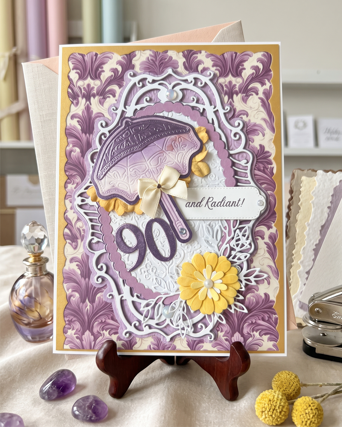 A handmade 90th birthday card with a purple damask pattern, white filigree, a lavender umbrella, yellow paper flower, and a cream ribbon bow, displayed on a small wooden stand. Made by Del Cards NZ.