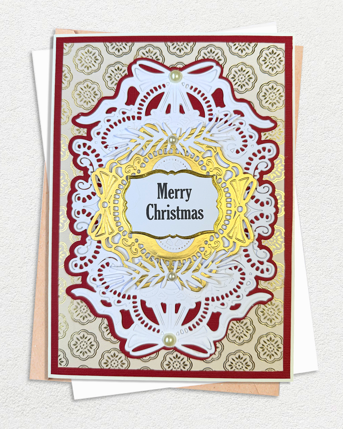 An ornate Christmas greeting card with layered white lace-like cutout details, a gold central frame, and the words “Merry Christmas” in black text. The card is layered over a beige patterned cardstock and a white envelope, photographed on a light textured background.
