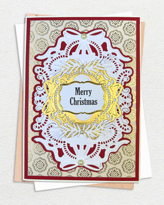 An ornate Christmas greeting card with layered white lace-like cutout details, a gold central frame, and the words “Merry Christmas” in black text. The card is layered over a beige patterned cardstock and a white envelope, photographed on a light textured background.
