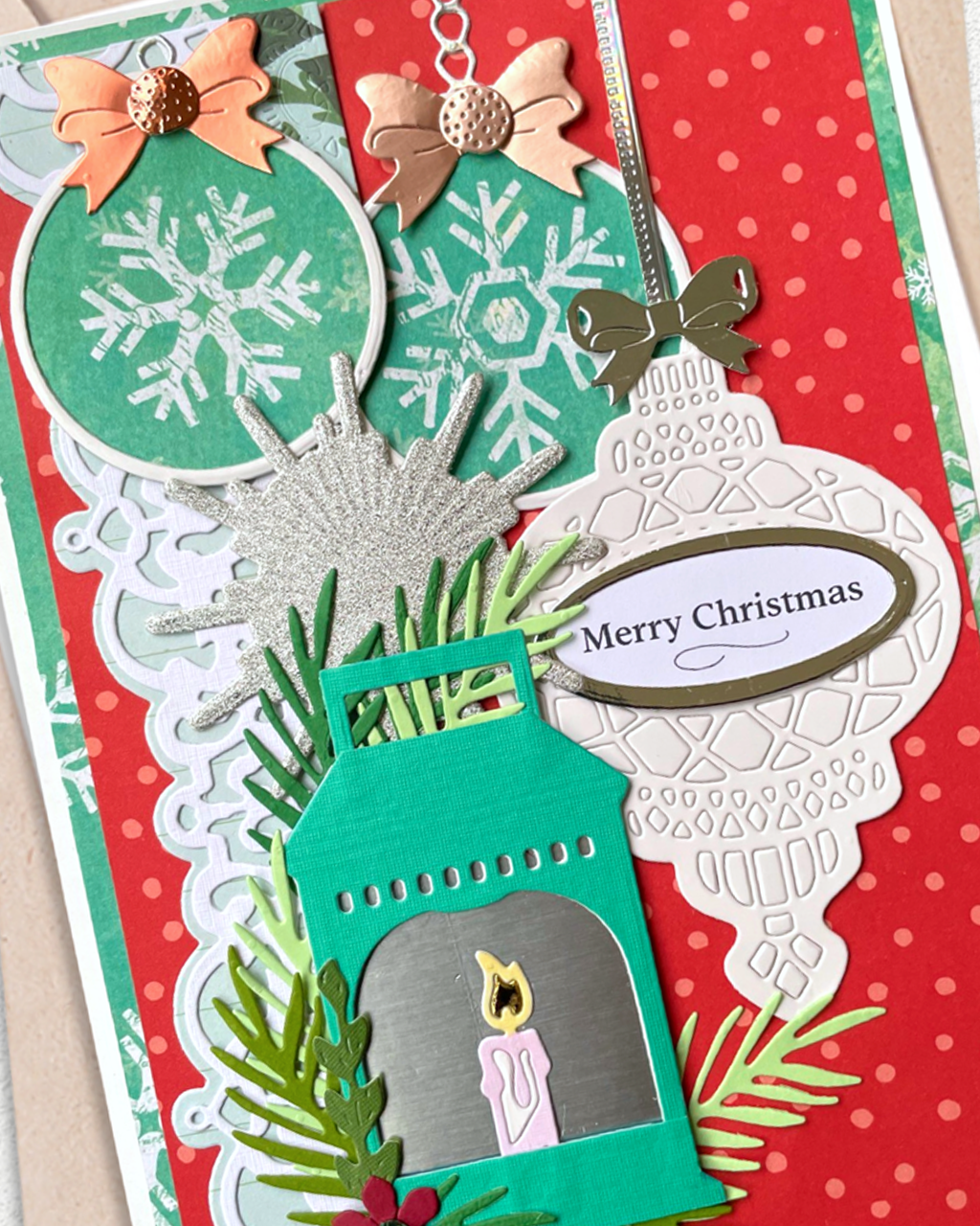 A close-up of a handmade Christmas card featuring layered paper ornaments, a teal lantern with a candle, green pine branches, and a white scalloped ornament tag that reads “Merry Christmas.”.
