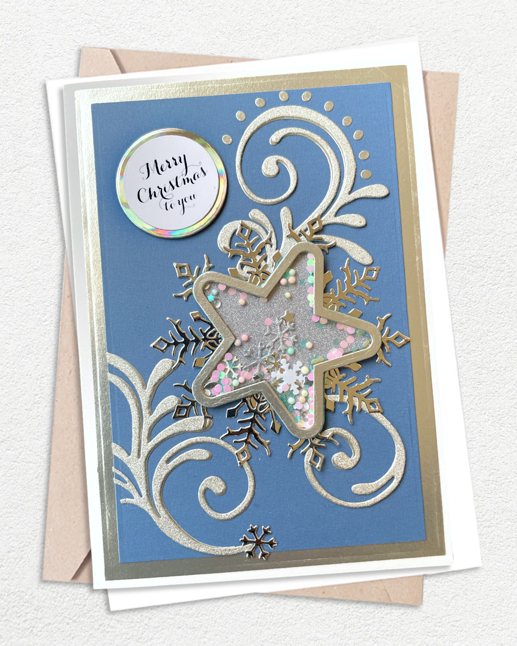 A square Christmas greeting card with a blue background, silver and gold embossed swirls, a large star-shaped cutout filled with glitter and small pink and white confetti, and a round holographic sticker that reads 'Merry Christmas to you.' The card is shown with a kraft envelope on a light textured background.