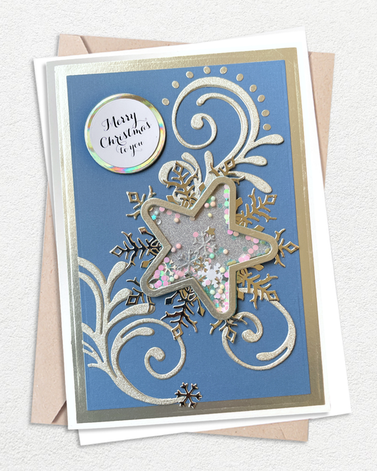 A square Christmas greeting card with a blue background, silver and gold embossed swirls, a large star-shaped cutout filled with glitter and small pink and white confetti, and a round holographic sticker that reads 'Merry Christmas to you.' The card is shown with a kraft envelope on a light textured background.
