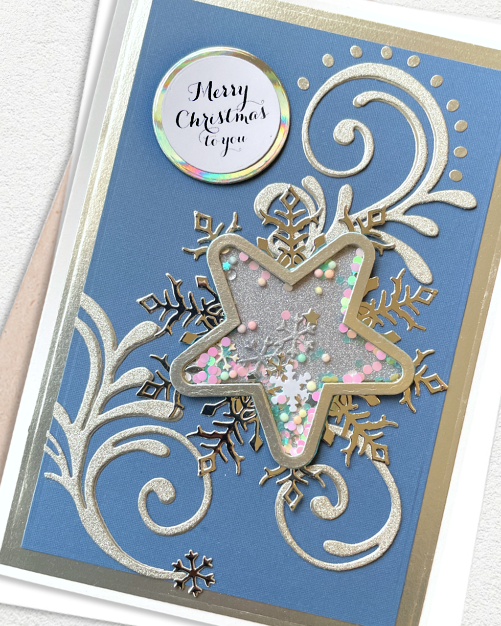 A close-up of a handmade Christmas greeting card featuring a blue cardstock background with silver glittery scrollwork, a large star-shaped cutout filled with pastel pink, green, and white glitter, gold glitter snowflakes, and a round holographic sticker that reads 'Merry Christmas to you'.