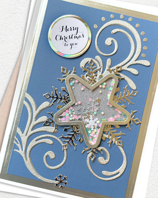 A close-up of a handmade Christmas greeting card featuring a blue cardstock background with silver glittery scrollwork, a large star-shaped cutout filled with pastel pink, green, and white glitter, gold glitter snowflakes, and a round holographic sticker that reads 'Merry Christmas to you'.