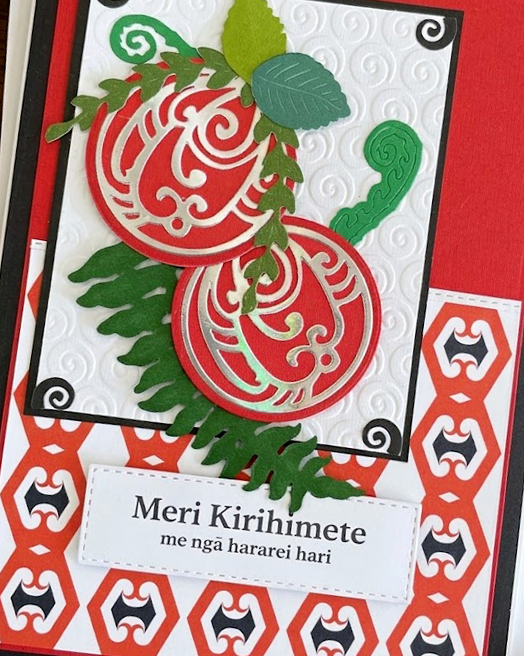 A Maori  Christmas card with intricate Māori patterns, featuring text 'Meri Kirihimete' and 'Merry Christmas and happy holidays' on a red background, with a green leaf and berry design.