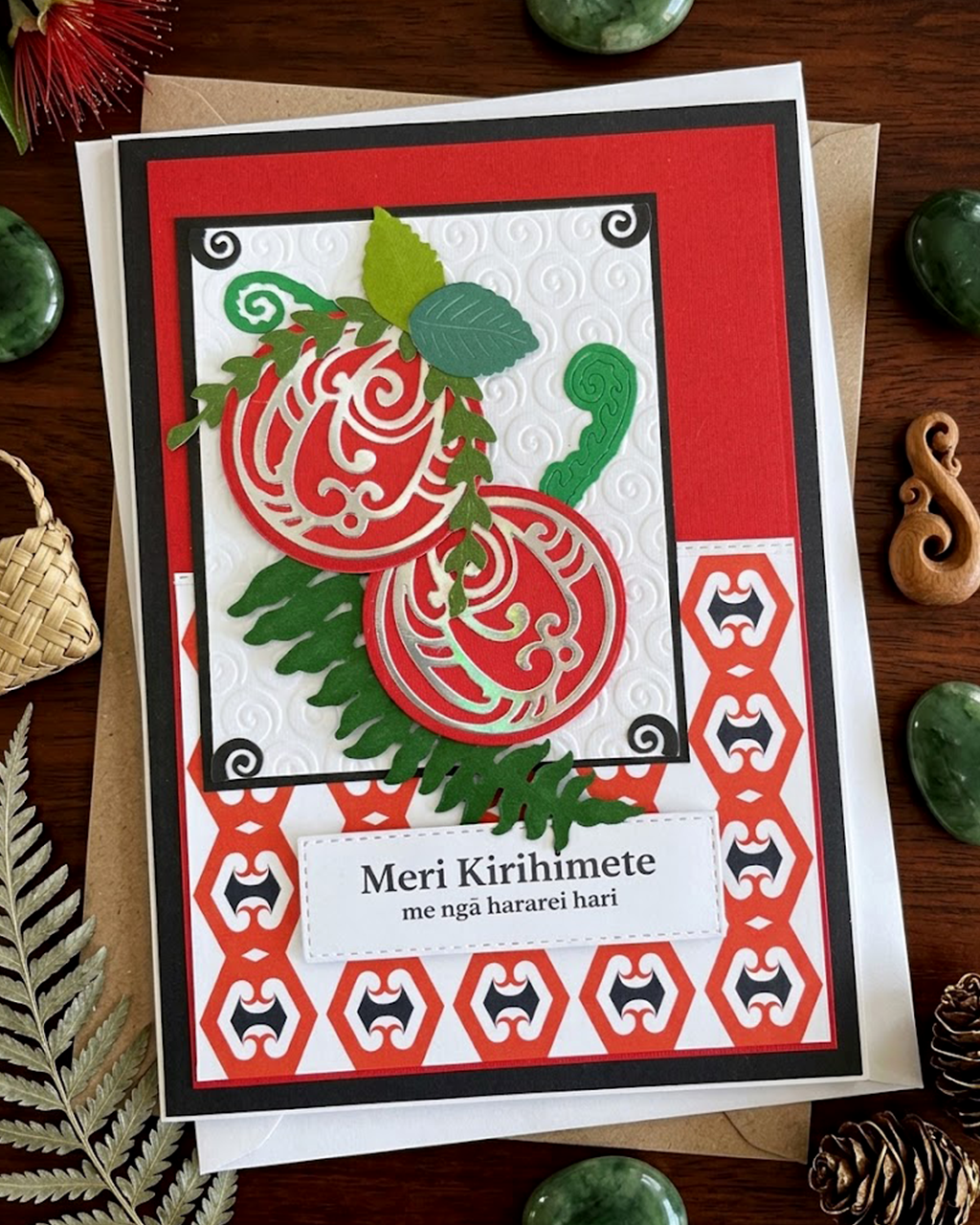 A MAori Christmas card with intricate Māori patterns, featuring text 'Meri Kirihimete' and 'Merry Christmas and happy holidays' on a red background, with a green leaf and berry design, made in NZ by Del Cards.