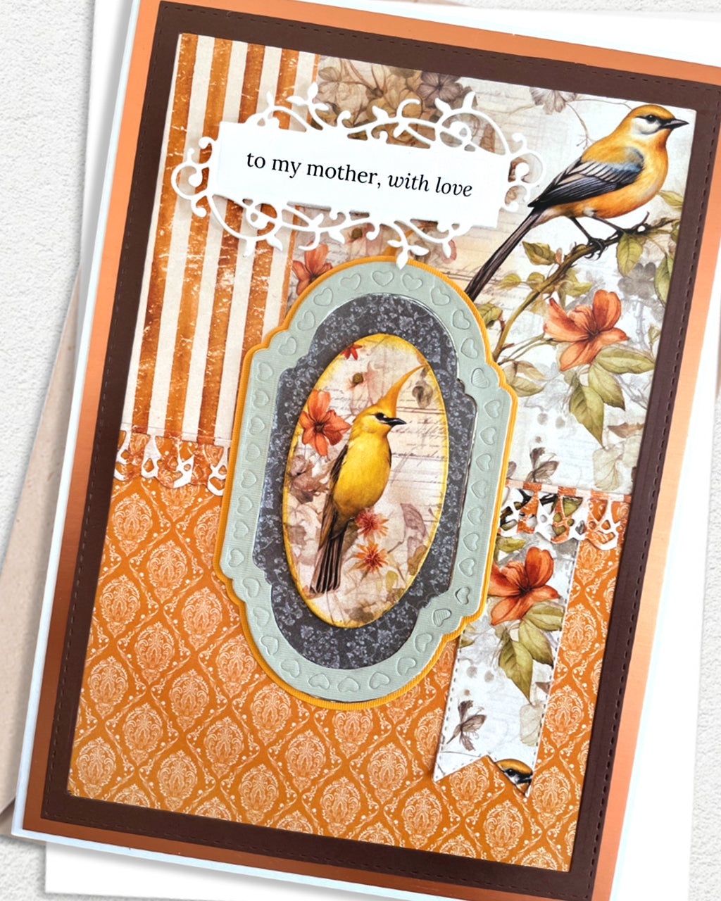 Close-up detail of a handmade luxury mother's card showing the upper portion — golden songbird perched on a branch above the oval centrepiece, with autumn botanicals and orange damask patterned paper. Sentiment reads "to my mother, with love." Handmade in in New Zealand by Del Cards NZ.