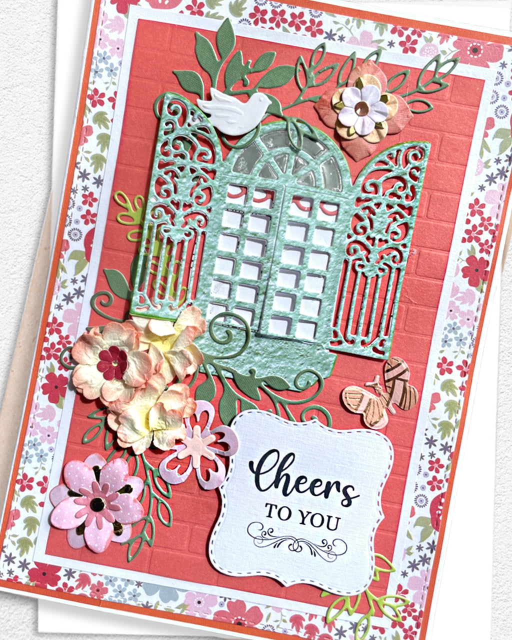 Close-up of a handmade luxury card showing an ornate teal filigree garden gate die-cut with a perched bird, surrounded by hand-applied pink and cream paper flowers, against a coral red background with floral border paper. Sentiment reads "Cheers TO YOU." Handmade greeting card made in New Zealand.