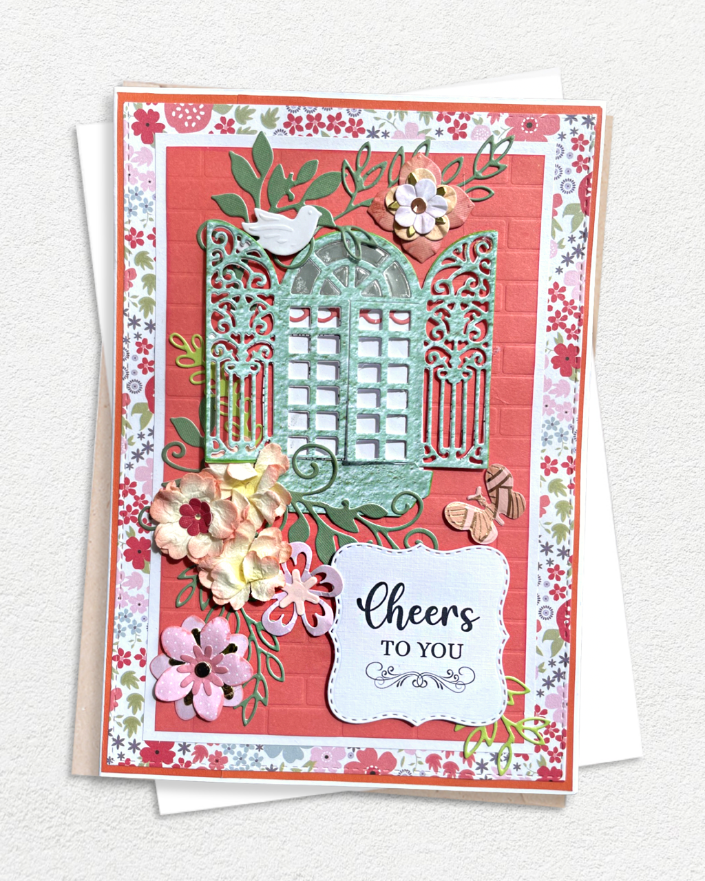 Handmade luxury greeting card with a warm coral red textured cardstock base, bordered by a delicate ditsy floral patterned paper in pink, white and green. A large ornate teal die-cut garden gate with filigree lattice detail and scrollwork dominates the centre, with a small bird perched at the top. Hand-applied paper flowers in pink, cream and red cluster around the gate. A white tag-style sentiment banner at the base reads "Cheers to You" in bold and script lettering. Handmade in NZ by Del Cards NZ.