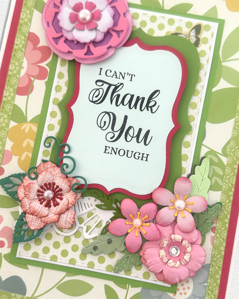 A close-up of a handmade layered card featuring the text 'I CAN'T Thank You ENOUGH' in black script on a pale mint label. The card is decorated with pink and peach paper flowers, green leaves, scalloped pink and green borders, and a butterfly embellishment.