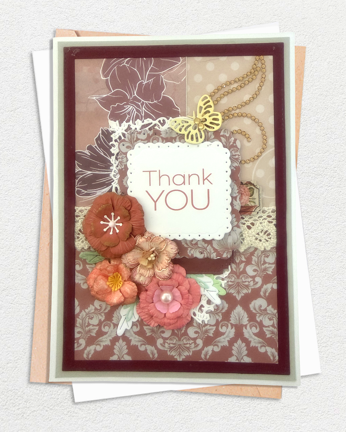 A handmade layered card with a 'Thank You' message, decorated with fabric flowers, a gold butterfly, lace, and damask patterns in earthy tones, displayed on top of envelopes against a textured white background, made by Del cards NZ.