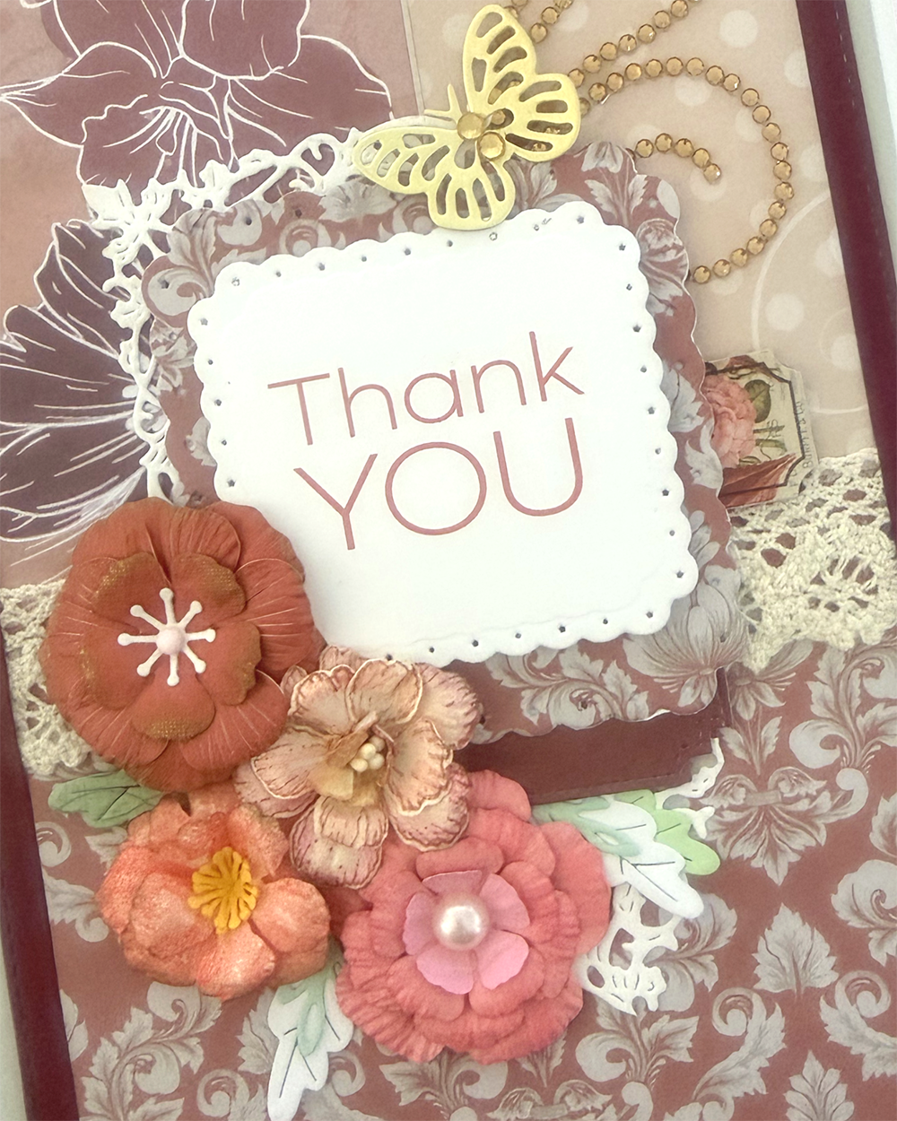 A close-up of a decorative handmade card or scrapbook page with layered floral embellishments, lace, a butterfly embellishment, and a central white scalloped label that reads “Thank You” in pink lettering.