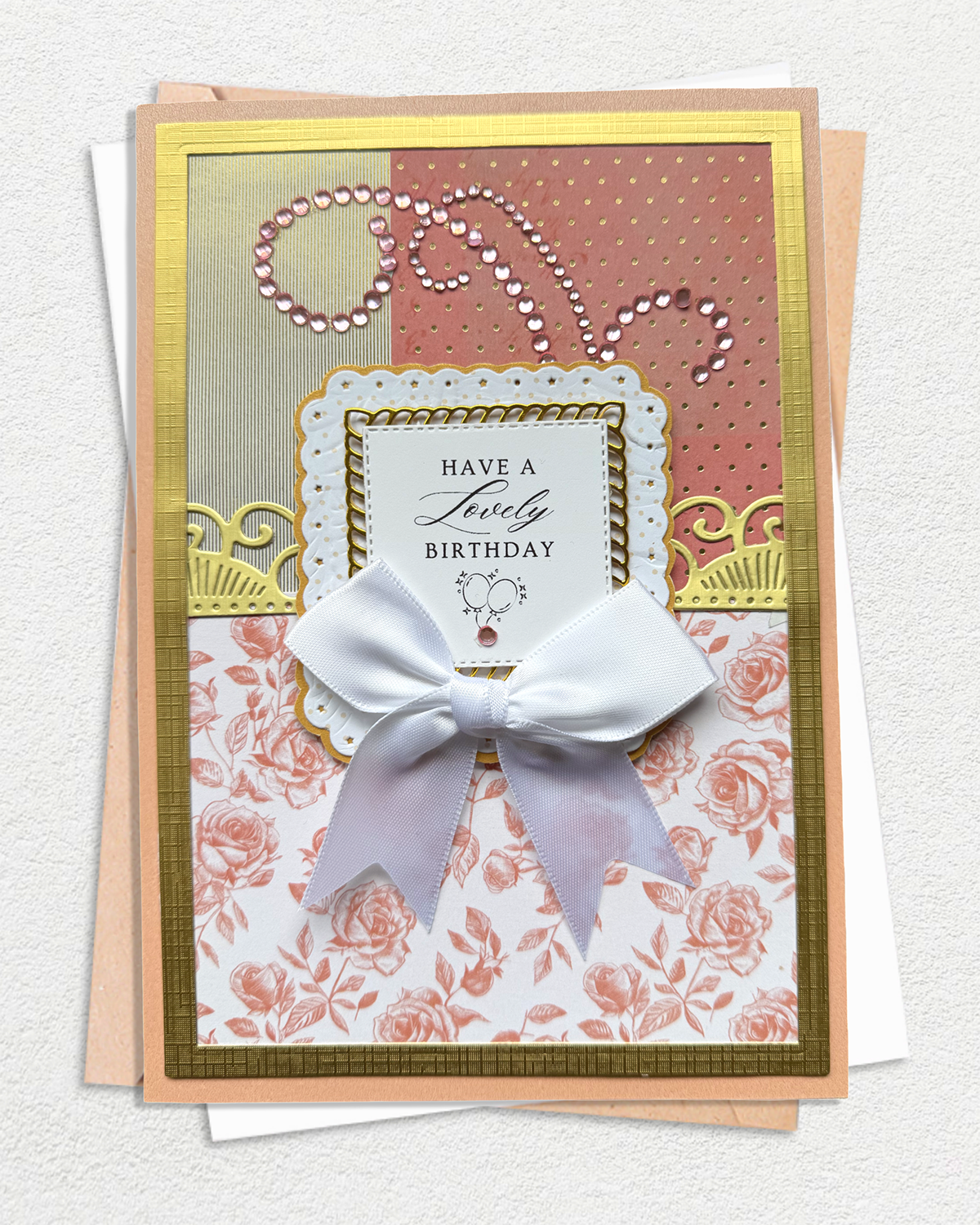 An elegant handmade birthday card with a large white bow, layered on top of a peach envelope. The card features a gold border and is decorated with multiple patterned sections: a gold glitter section, a dusty pink polka dot section with rhinestone accents, and a pink rose print section. Made by Del Cards NZ.