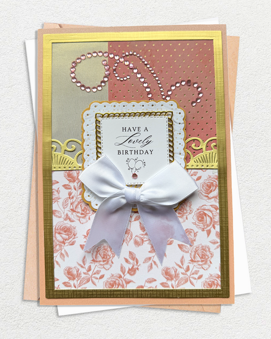 An elegant handmade birthday card with a large white bow, layered on top of a peach envelope. The card features a gold border and is decorated with multiple patterned sections: a gold glitter section, a dusty pink polka dot section with rhinestone accents, and a pink rose print section. Made by Del Cards NZ.