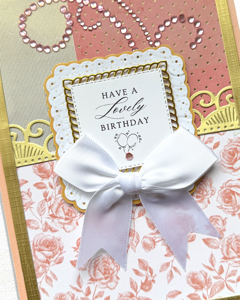 A close-up of a handmade birthday card featuring a white satin bow, layered white and pink floral paper, gold foil accents, and a central white card that reads 'HAVE A Lovely BIRTHDAY' with a small heart design.