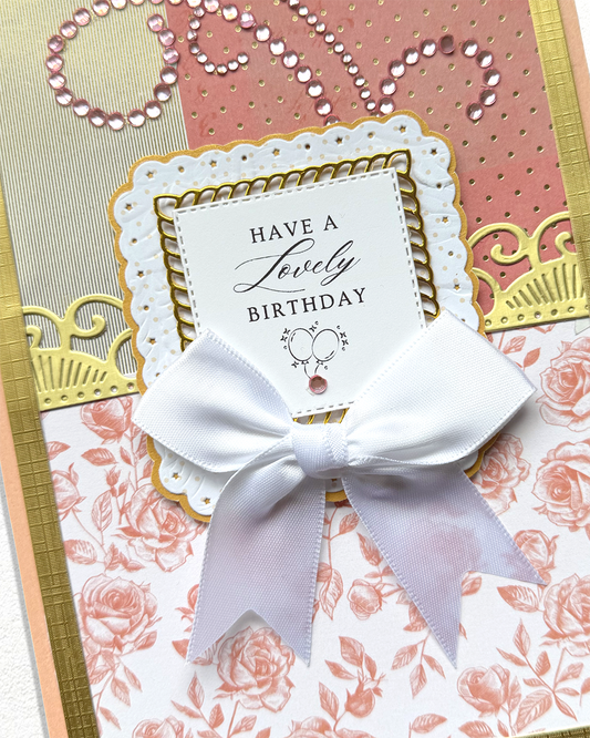 A close-up of a handmade birthday card featuring a white satin bow, layered white and pink floral paper, gold foil accents, and a central white card that reads 'HAVE A Lovely BIRTHDAY' with a small heart design.