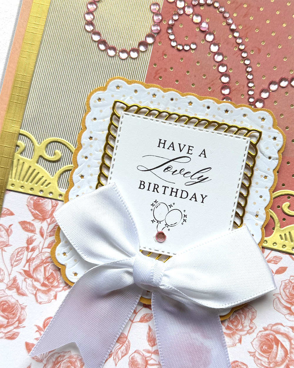 A close-up of a handmade birthday card featuring a white satin bow, a scalloped white paper label that reads 'HAVE A Lovely BIRTHDAY,' and layered floral and metallic pink, gold, and white cardstock backgrounds.