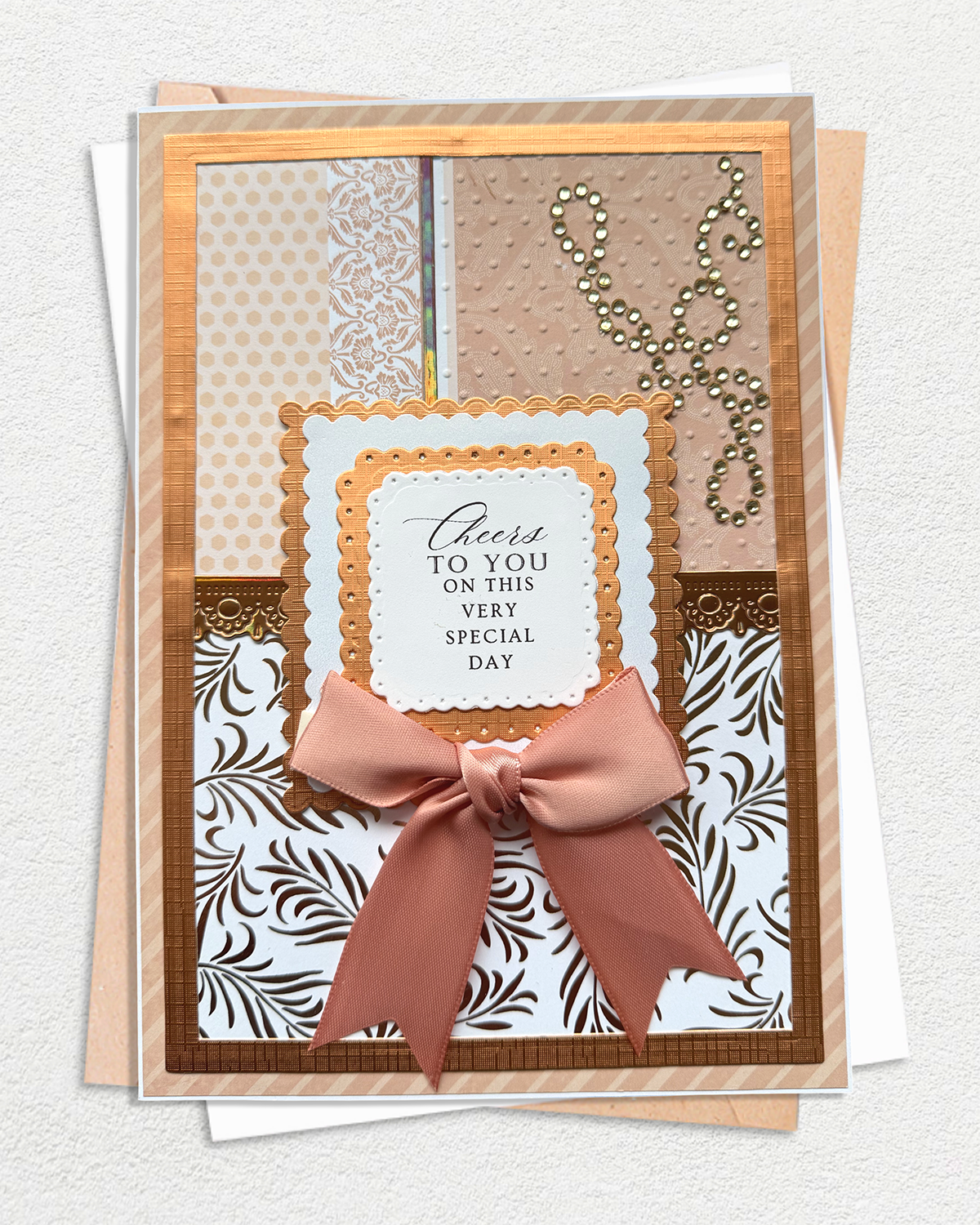 A handmade greeting card with a layered design, featuring a dusty pink satin bow, patterned paper sections, and a central white label that reads 'Cheers to you on this very special day'.
