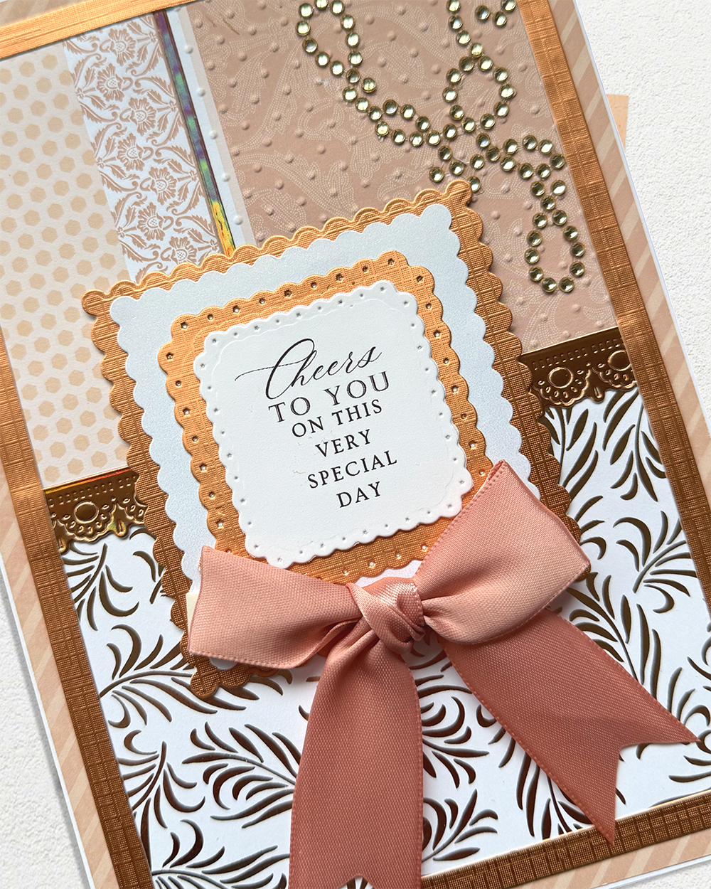 A close-up of a handmade greeting card featuring a peach satin bow, layered scalloped borders, patterned paper, and a central white card that reads, 'Cheers TO YOU ON THIS VERY SPECIAL DAY.'.