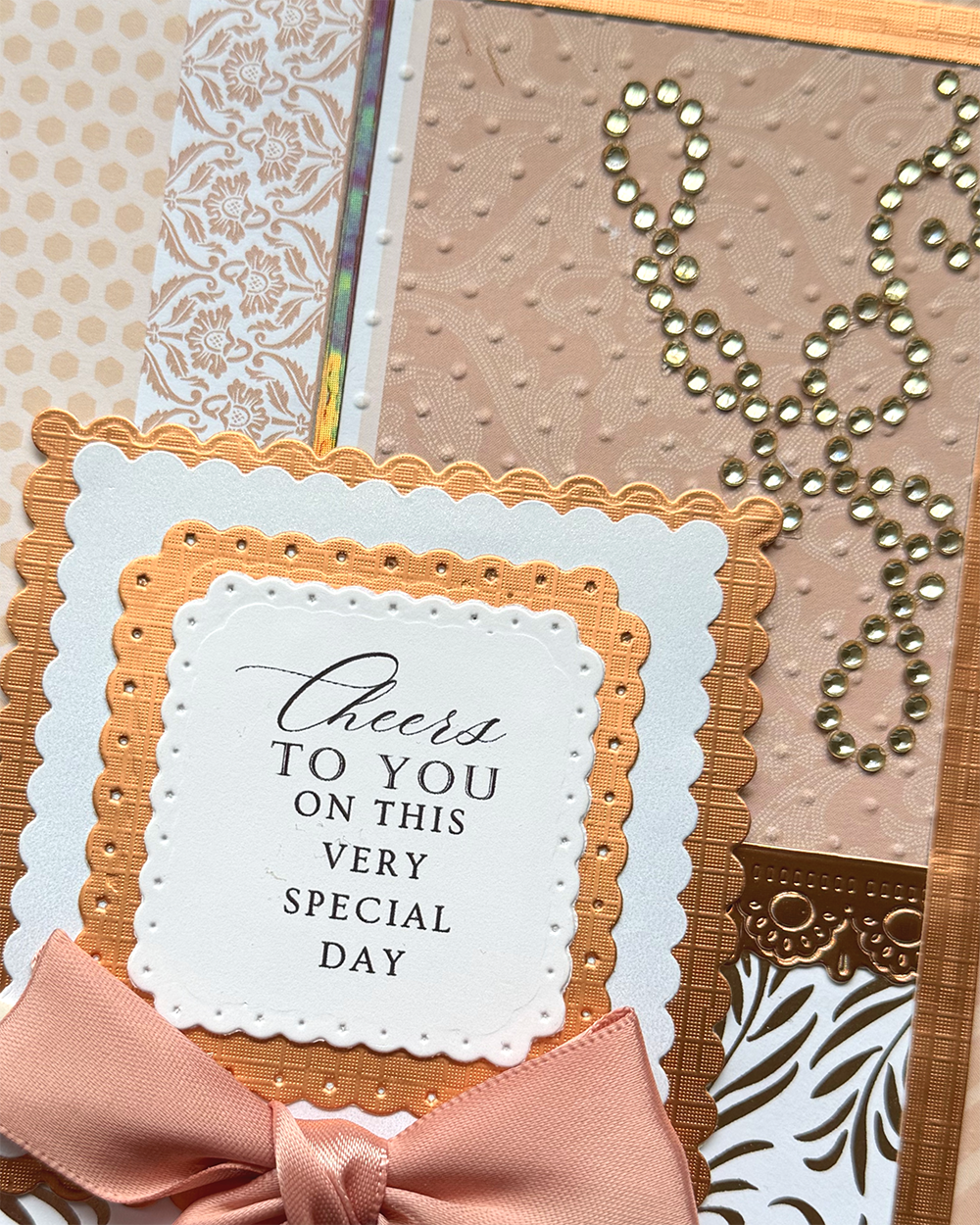A close-up of a handmade greeting card featuring layered scalloped borders in white, gold, and peach, a peach satin bow, and a central white card that reads 'Cheers TO YOU ON THIS VERY SPECIAL DAY'. The card includes textured beige panels, metallic gold rhinestone embellishments, and patterned paper accents.