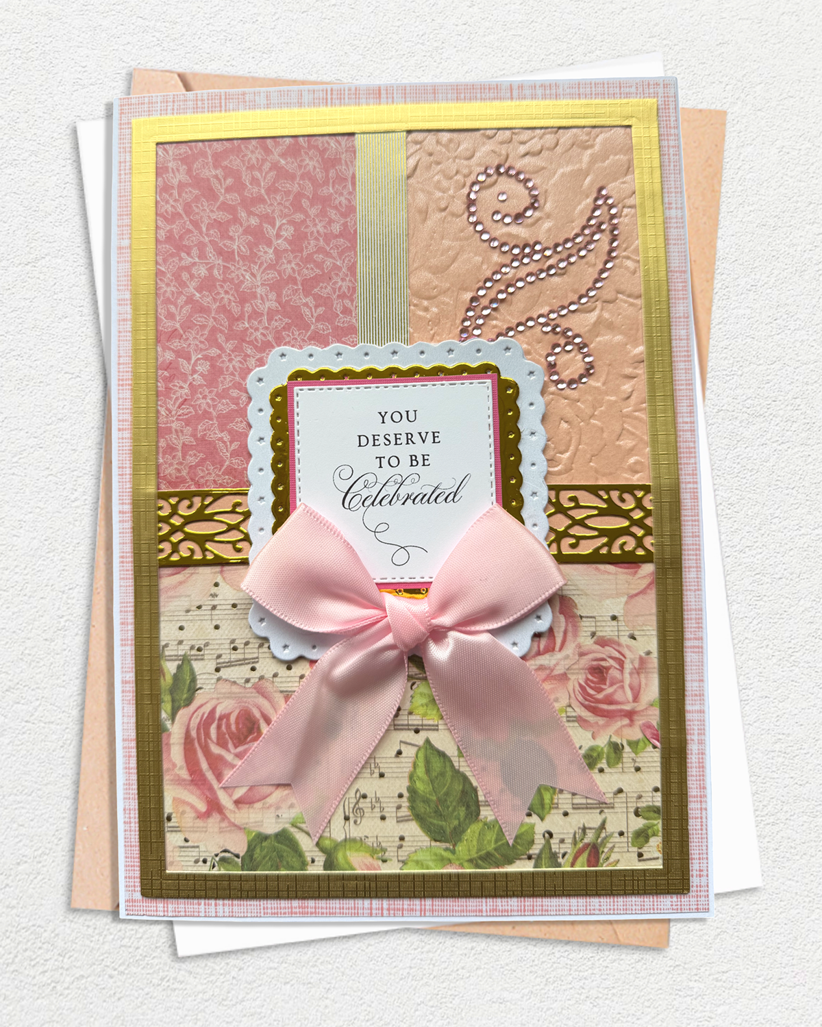 A handmade layered greeting card with a large pink satin bow, featuring floral and patterned paper sections, gold borders, and the text 'YOU DESERVE TO BE Celebrated'.