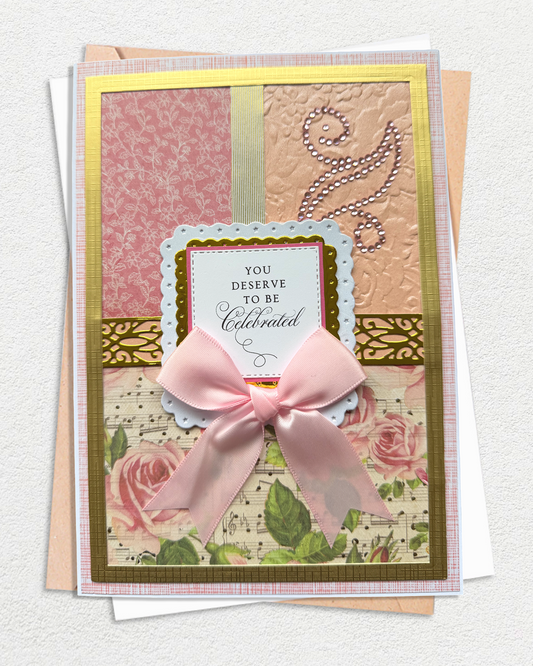 A handmade layered greeting card with a large pink satin bow, featuring floral and patterned paper sections, gold borders, and the text 'YOU DESERVE TO BE Celebrated'.