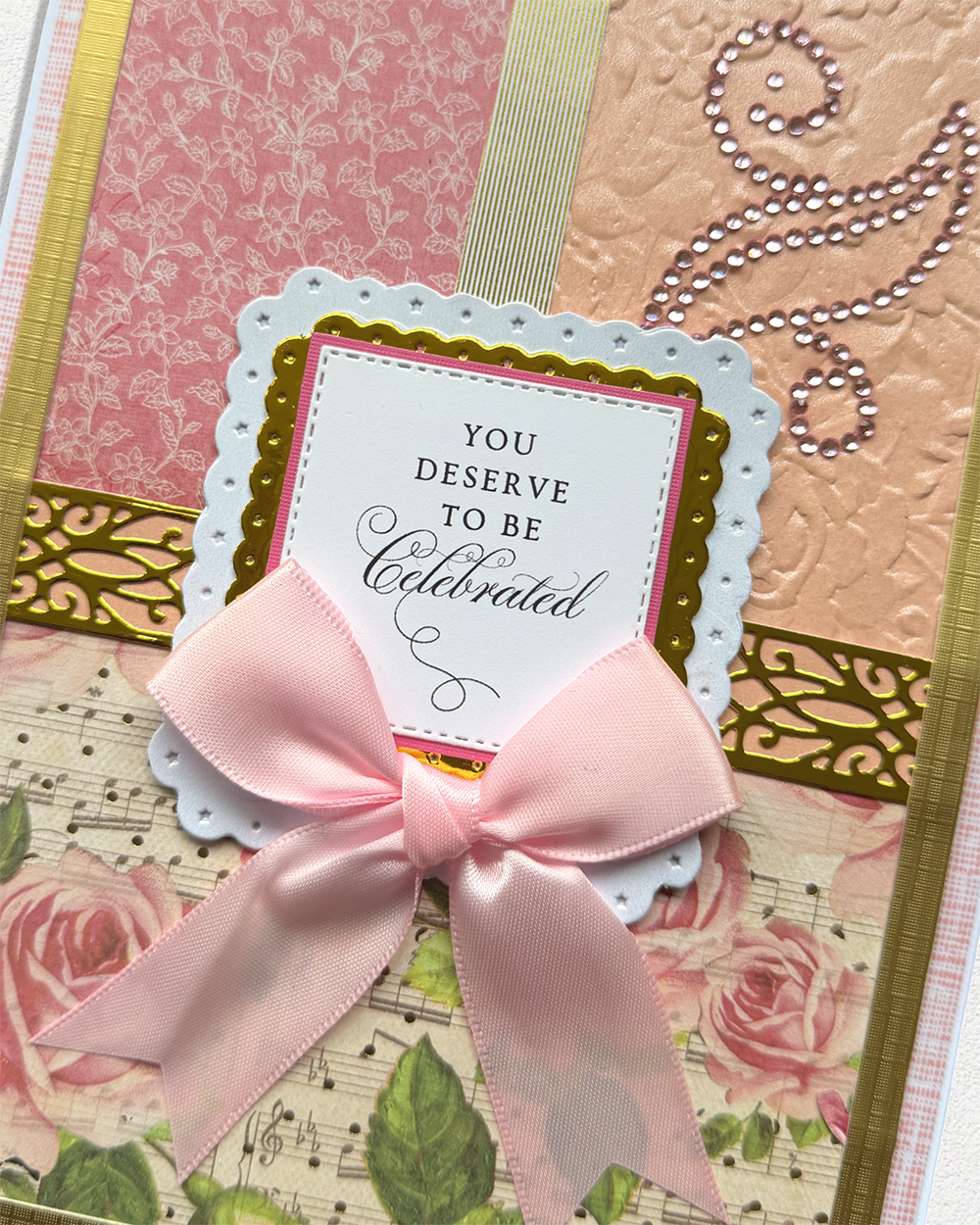A close-up of a decorative handmade greeting card featuring a pale pink satin bow, scalloped white and pink cardstock layers, floral print details, and the printed phrase 'YOU DESERVE TO BE Celebrated'.