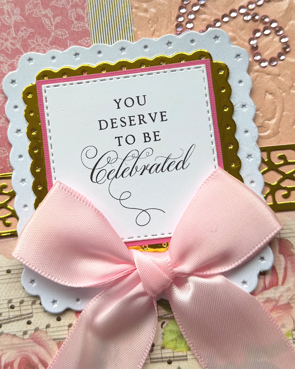 A close-up of a handmade greeting card or scrapbook paper design featuring a white scalloped card with the text 'YOU DESERVE TO BE Celebrated,' layered over pink and gold borders, a large pale pink ribbon bow, and decorative patterned paper in the background.