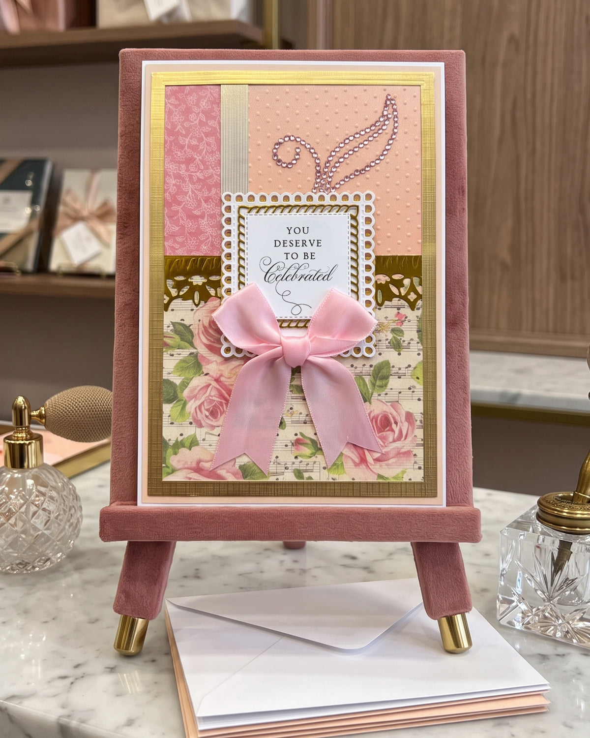 A handmade greeting card displayed on a pink velvet stand on a marble surface. The card features pink roses, a large pink bow, patterned pink and peach paper, gold foil borders, and a central white label that reads 'YOU DESERVE TO BE Celebrated' - made in NZ by Del Cards NZ.