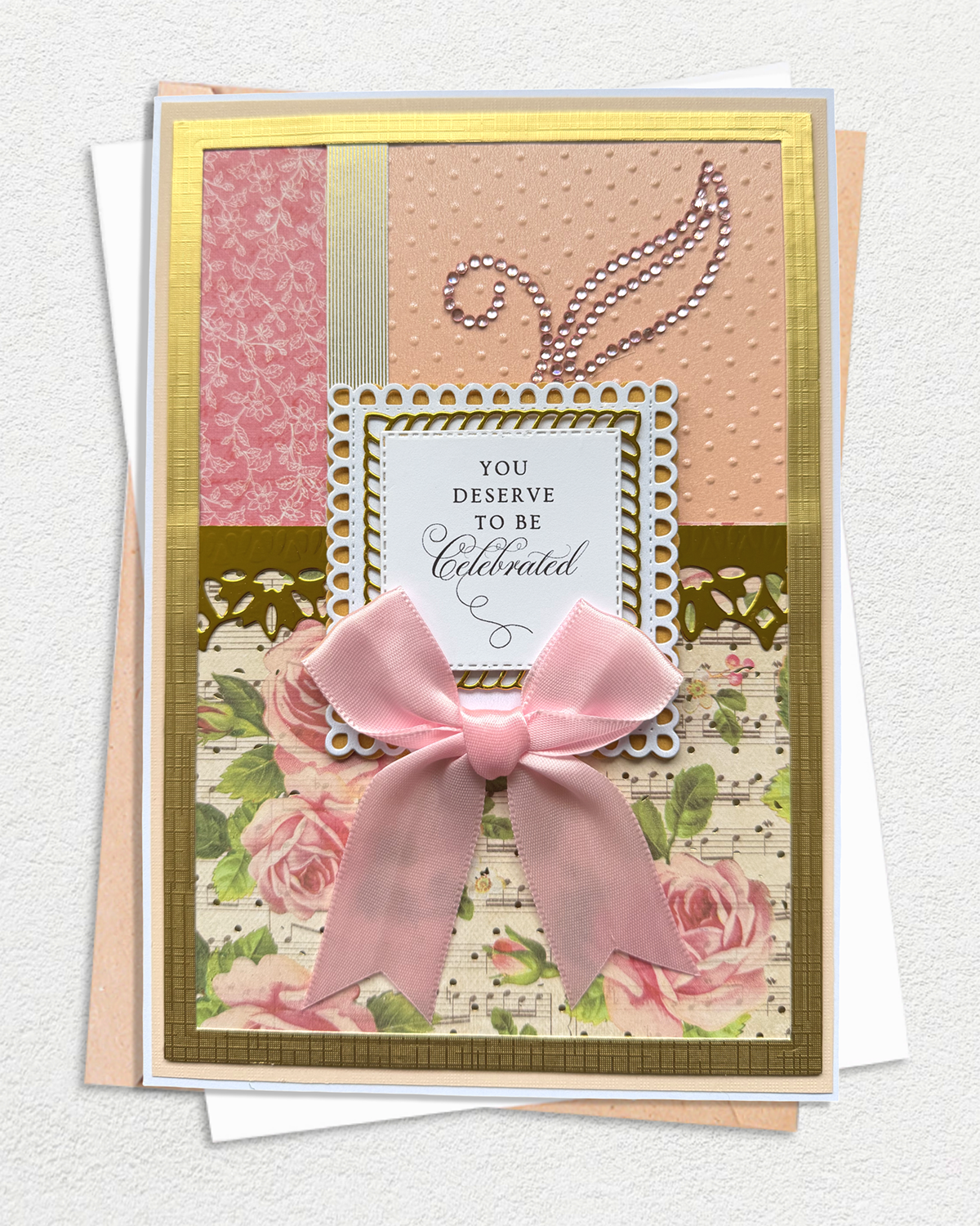 A handmade layered greeting card with a large pink satin bow, featuring floral and musical note patterns, gold borders, and a central white label that reads 'YOU DESERVE TO BE Celebrated'.