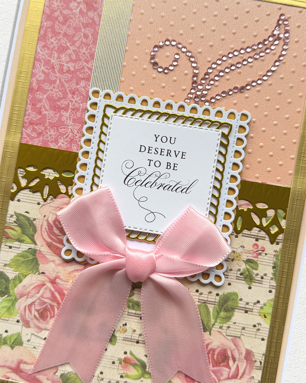 A close-up of a handmade greeting card or scrapbook page featuring a pink satin bow, layered floral and patterned paper sections, and a central white card that reads, 'YOU DESERVE TO BE Celebrated.'.