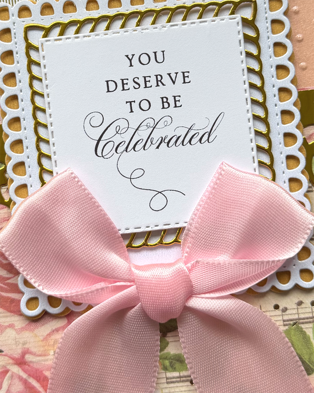 A close-up of a handmade greeting card or invitation featuring a large pink satin bow, a white central card with the text 'YOU DESERVE TO BE Celebrated,' and layered white and gold scalloped borders over a floral background.