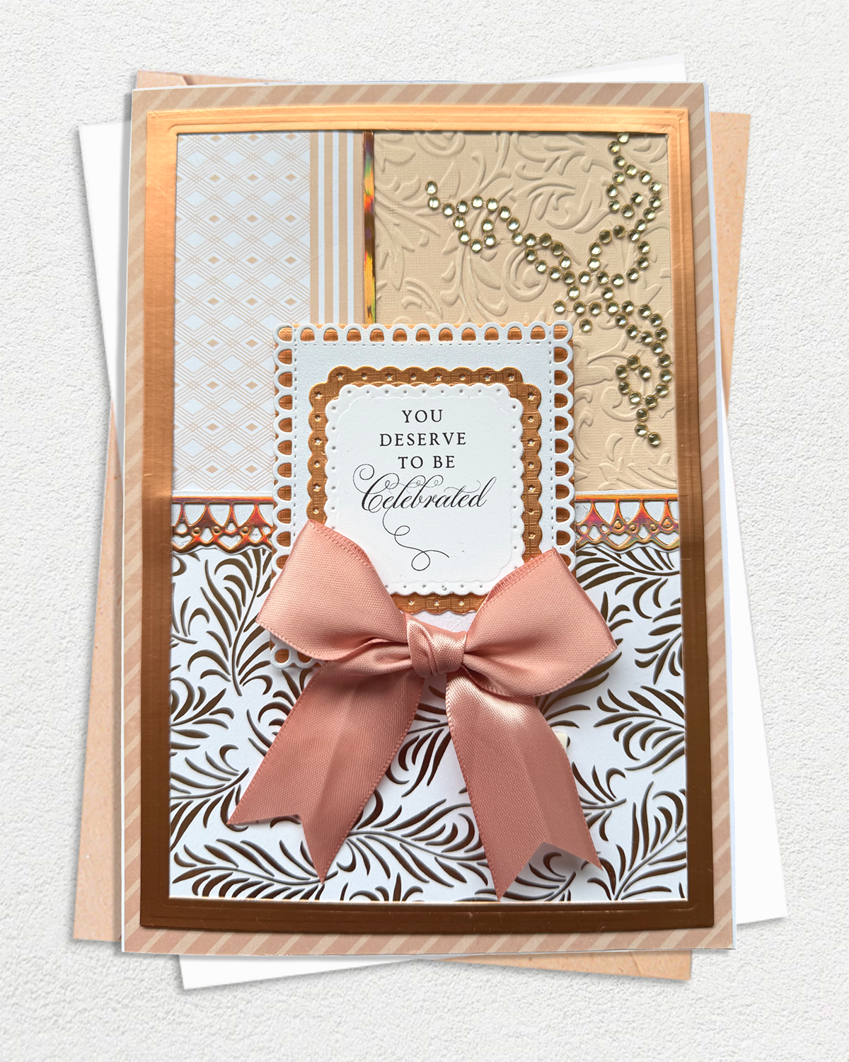 A handmade greeting card with a large dusty pink satin bow, layered over patterned paper and a white envelope, displayed on a textured light gray background.