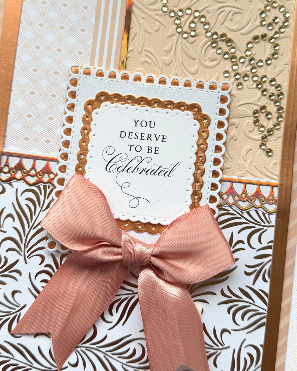 A close-up of a decorative handmade greeting card or invitation featuring layered scalloped paper borders, a peach satin bow, and the printed message 'YOU DESERVE TO BE Celebrated' on a patterned background.