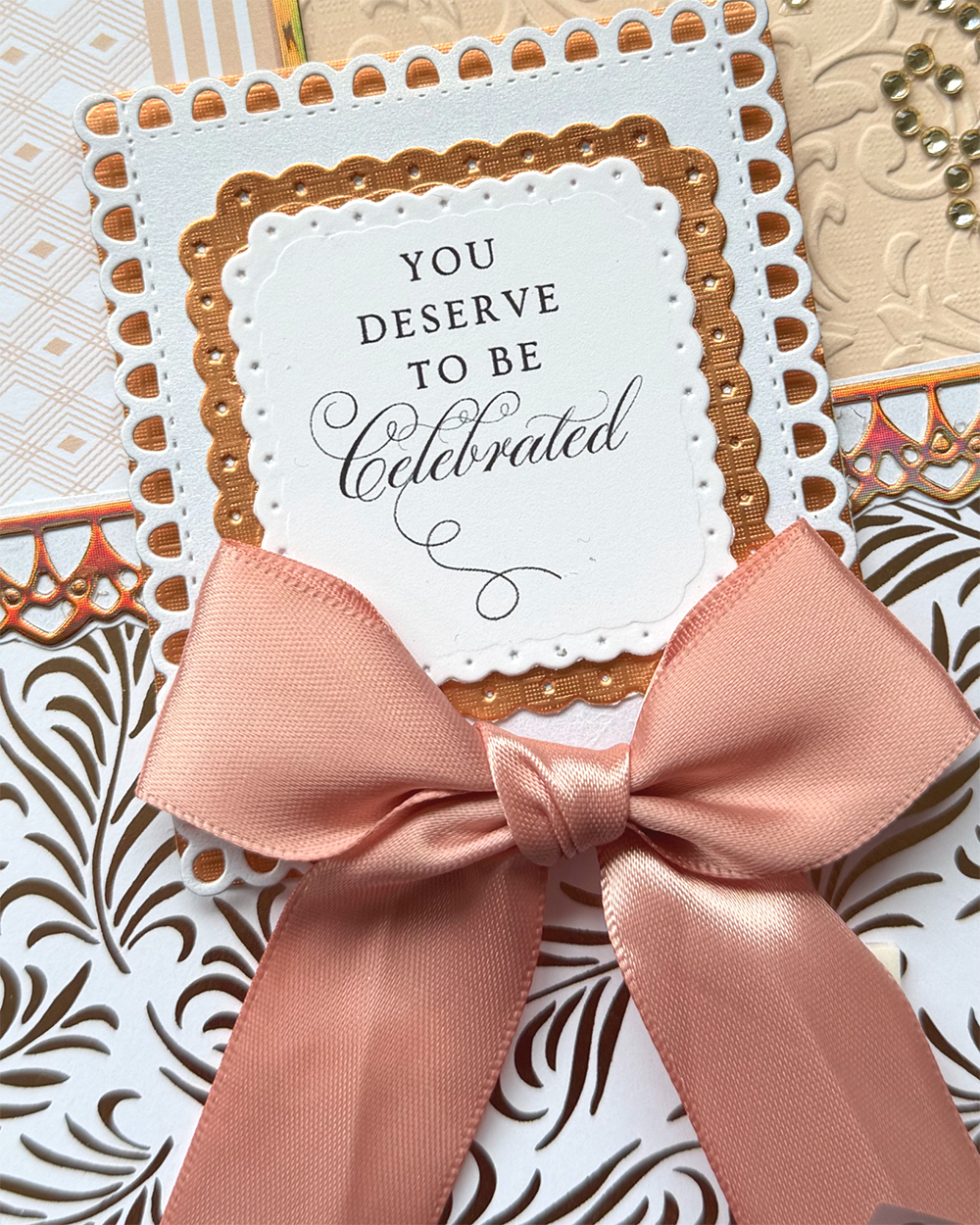 A close-up of a handmade greeting card featuring a large satin pink ribbon bow, layered white and gold paper borders, and the printed message 'YOU DESERVE TO BE celebrated'.